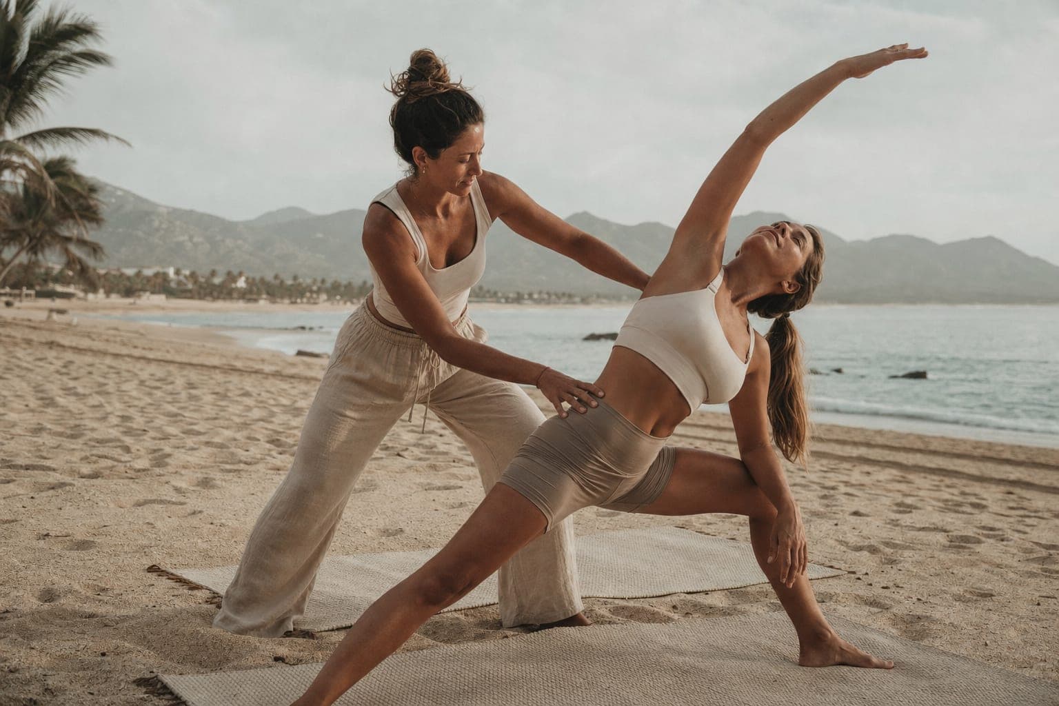 A coach guiding a student through a stretch on the sand at sunrise